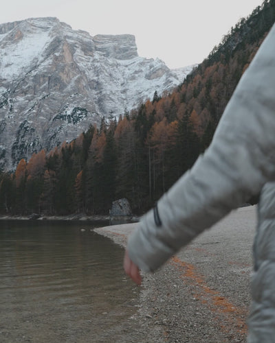 A woman is wearing a light grey light down coat and walking by the shoreline. There are mountains behind her.