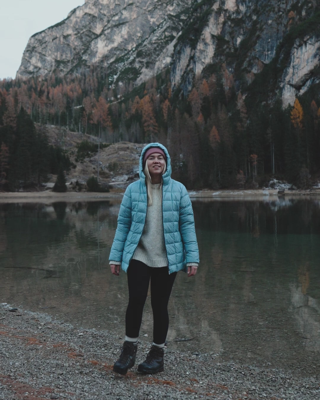 A woman is walking by a lake wearing a light blue light down coat and lilac beanie.